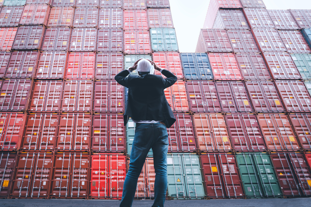 Worker or foreman standing in front of stacked cargo containers and hands hold his head wears white hardhat and hold face mask ack like a shock or surprise. Difficulties in transport