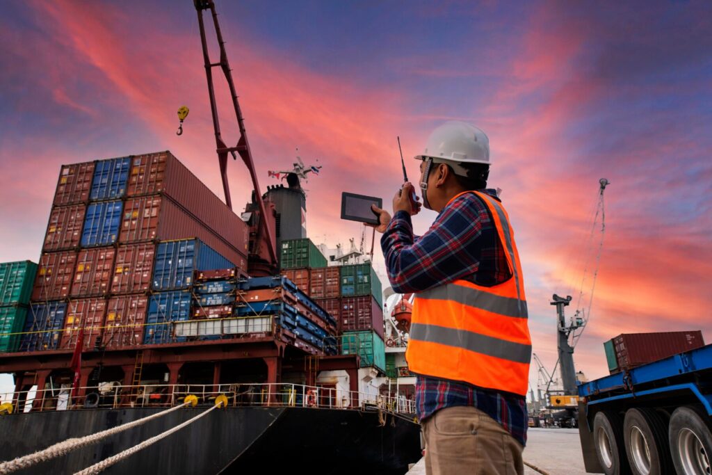 containers being moved around at shipping port. Worker wearing hi-vis and helmet communicating over radio